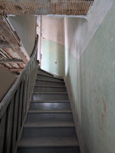 Altbau-Treppenhaus vor Sanierung Braune Treppe mit grauem Geländer, führt zu einem hellen, verputzten Flur.